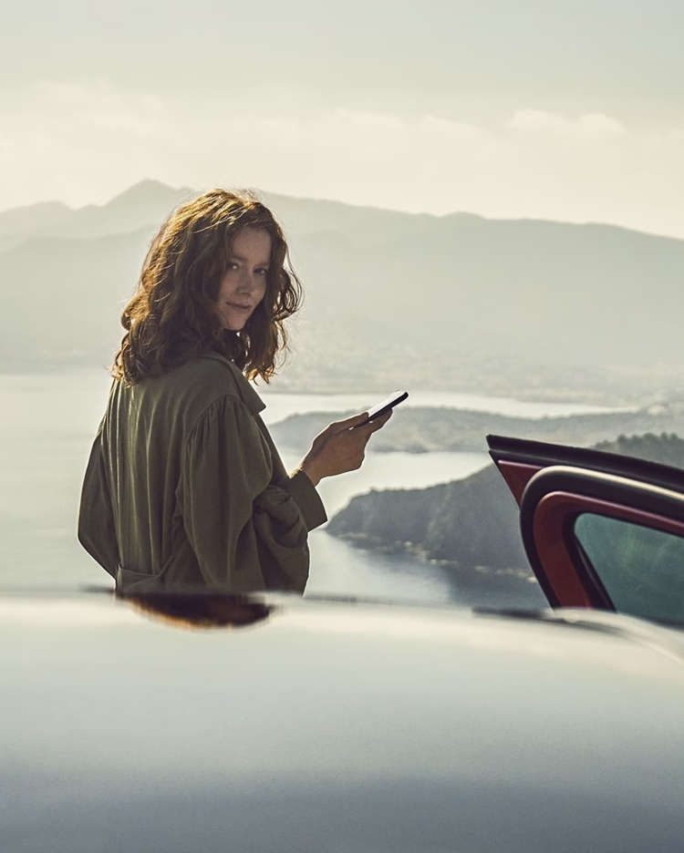 Дівчина зі смартфоном стоїть за автомобілем на фоні морської затоки