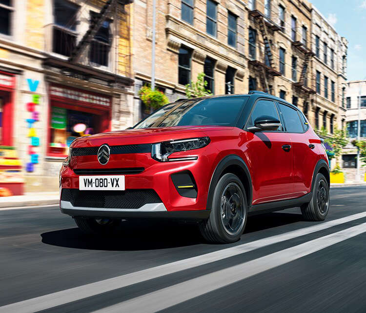 Новий Citroën ë-C3 червоного кольору їде по вулиці, вигляд спереду з лівого боку